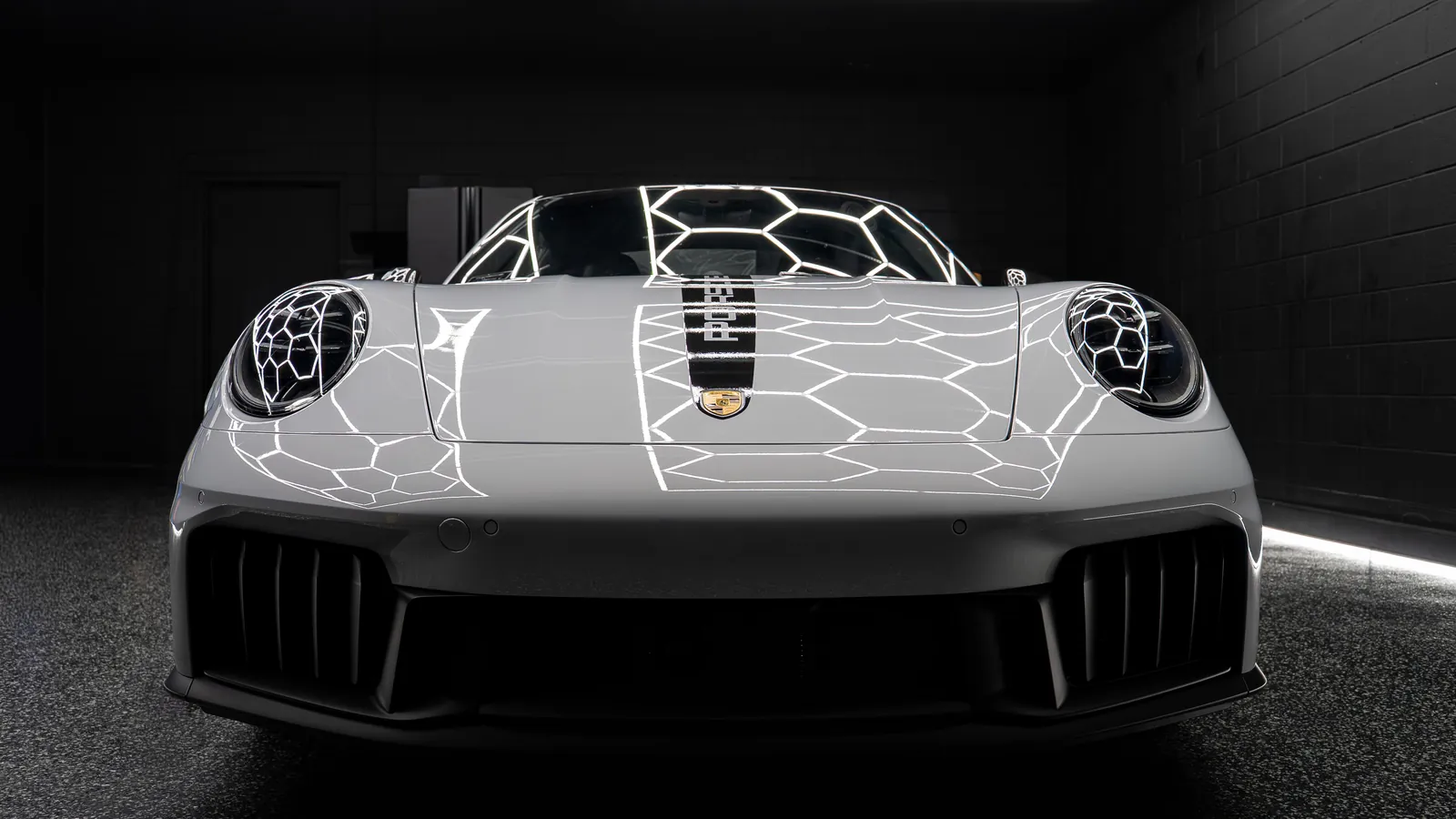 Front of a light grey Porsche 911 Carrera GTS with the black hood stripe and neon ceiling light reflected in the paint
